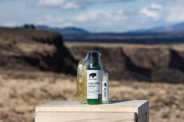 A bottle and a jar of "sage pine lotion" placed on a wooden stand, set against a landscape of open plains and distant hills under a clear sky.