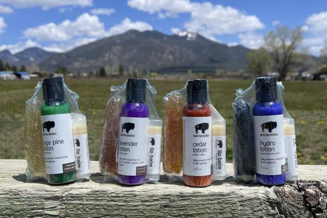 A row of six lip balm products displayed on a weathered wooden table, with scenic mountains and a blue sky with clouds in the background.