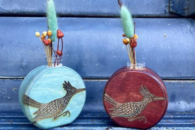 Two oval-shaped wooden vases are displayed on a textured background. Each feature a detailed roadrunner design on the front, both vases contain decorative dried elements, including colorful flower buds and fruits.