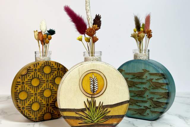 Three decorative wooden vases with dried flowers on a marble surface. From left to right, the first diffuser has an abstract pattern, the second features a sun and wheat design, and the third showcases a textured, peeling paint effect.