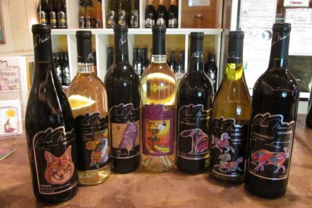 A collection of eight Black Mesa Winery bottles displayed on a wooden counter, featuring unique labels with colorful artistic illustrations of animals in various styles.