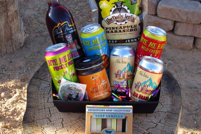 A wooden surface displays a collection of New Mexico-themed items, including bottled wine, a jar of salsa, beef jerky, assorted canned beverages with vivid labels, and a soap sampler set in a box labeled "New Mexico Collection.