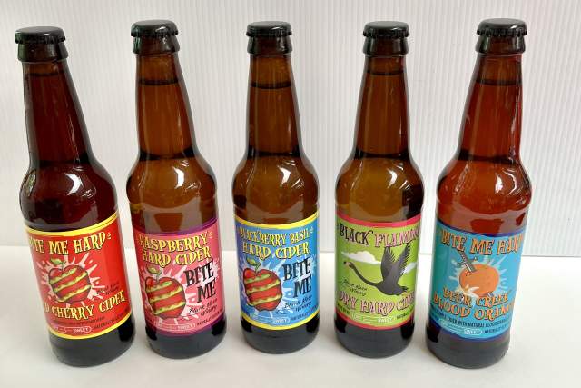 Five bottles of "Bite Me Hard" cider in various flavors displayed in a row against a white background.