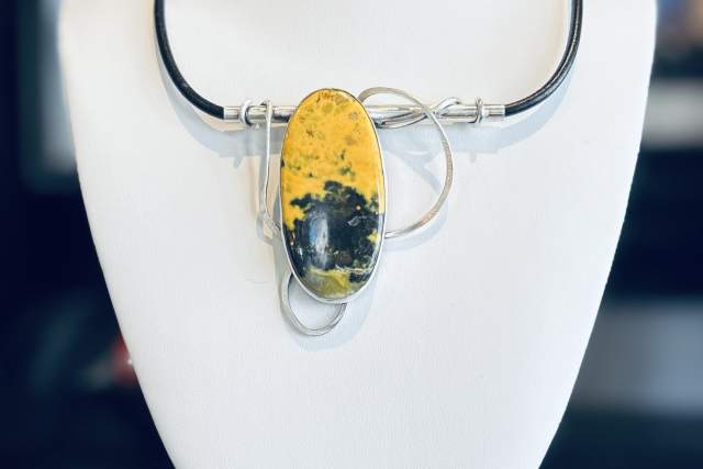 A necklace displayed on a stand featuring a large oval pendant with a vibrant yellow and black marbled design. The pendant is accentuated with intertwined metal loops and a sleek black cord. The background shows blurred framed images.