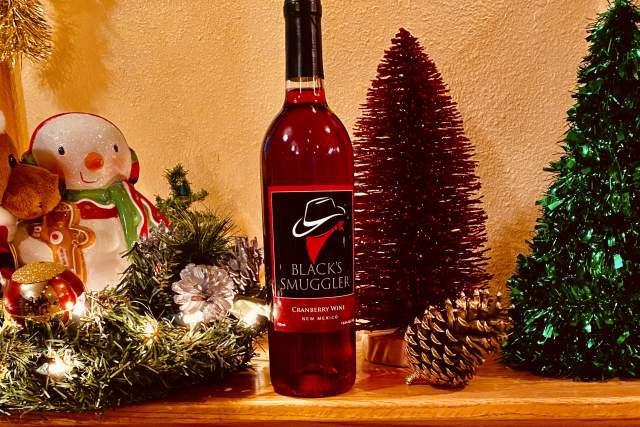 A bottle of Black's Smuggler Cabernet Wine placed on a mantel, flanked by a snowman decoration and two festive trees, one burgundy and one green, with string lights and a pine cone.