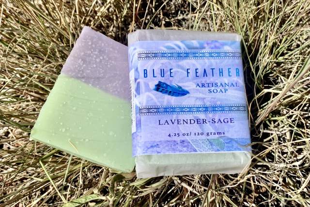 Two bars of Blue Feather Artisanal Soap, one unwrapped displaying a green and purple color, and the other wrapped with a lavender-sage label, set on a grassy surface.