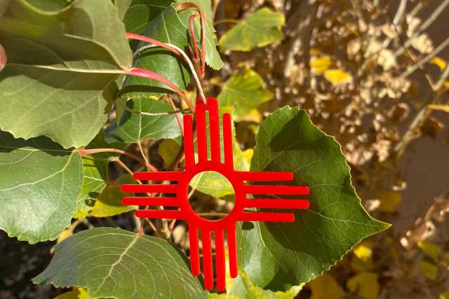 Close-up of the Zia sun symbol, vividly red, centered on a background of vibrant green and yellow leaves, set under a bright blue sky.