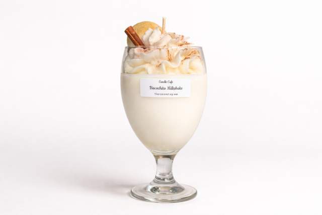 A candle in a glass topped with decorative whipped cream, a cinnamon stick, and a cookie on top.