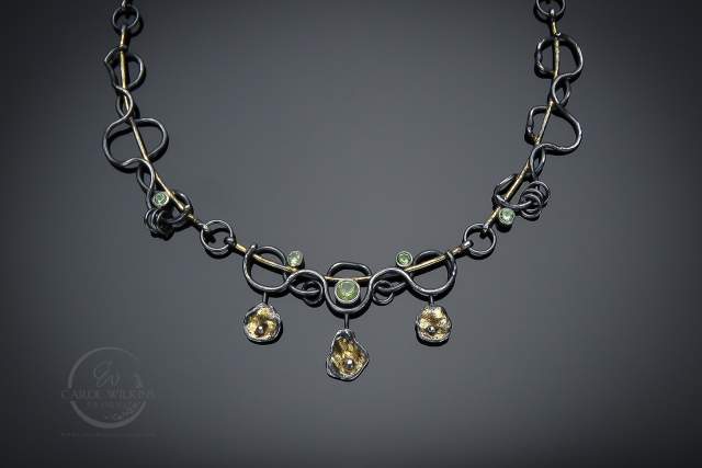 A necklace with intricate, dark metal loops and gold accents, adorned with gemstones and three hanging floral-shaped pendants, displayed on a dark background.