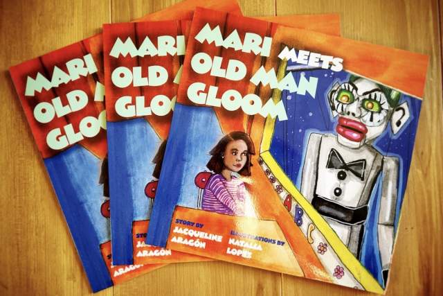 Four colorful book covers displayed on a wooden surface, titled "Mari Meets Old Man Gloom," written by Jacqueline Aragón and illustrated by Natzalie Loppli. Each cover features vibrant illustrations of characters, including a child and a whimsical figure of Zozobra.