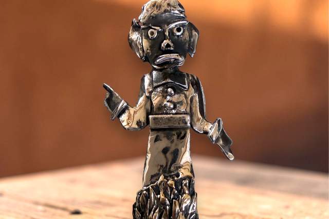 A metal figurine with an expressive face stands on a wooden surface. It features a rough, textured appearance with raised hands and is set against a warm, blurred background.