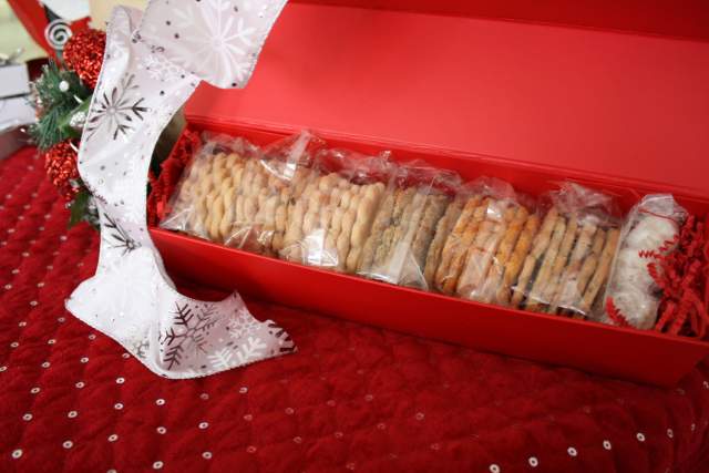 A red gift box filled with an assortment of cookies, each wrapped in clear plastic, placed on a red textured surface with a decorative white ribbon and holiday ornaments nearby.