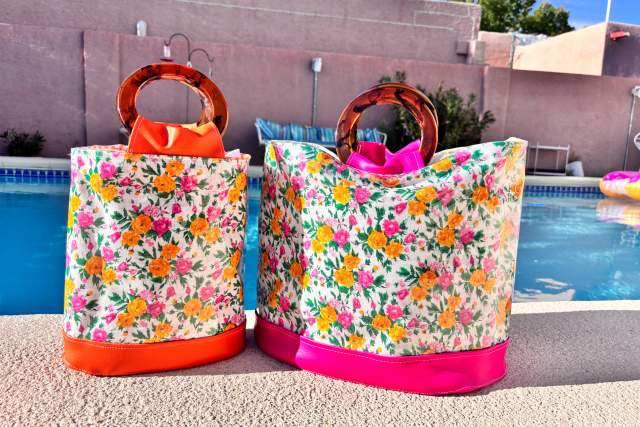 Two colorful floral patterned tote bags with bright handles, placed beside a pool with a clear blue sky above.