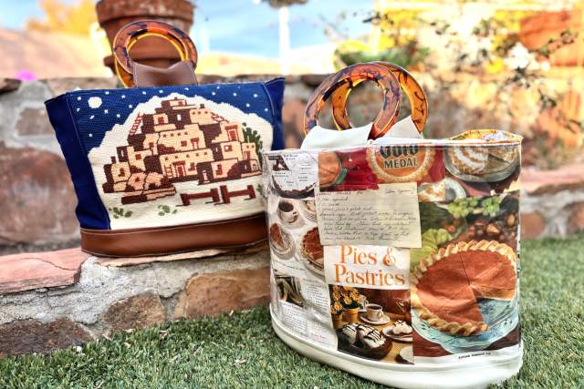 Two stylish tote bags sit on a grassy surface with a blurred garden in the background. The left bag features an adobe pueblo cross-stitched on denim, and the right bag showcases a vibrant collage of magazine clips related to pies and pastries. Both bags features additional small items peeking out.