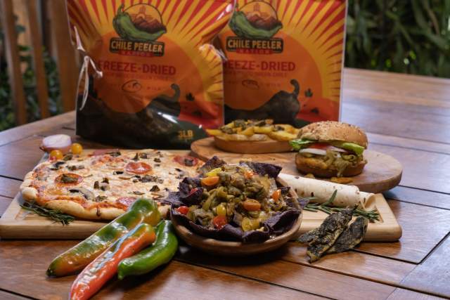 A variety of dishes featuring Chile Peeler brand products are displayed on a wooden table. In the background, two bags of Chile Peeler Freeze-Dried Green Chile are visible. The table includes a pizza topped with vegetables, a bowl of nachos with toppings, a hamburger, and an open burrito. Additionally, fresh green and red peppers are arranged on the table.