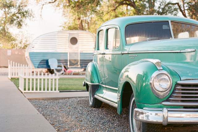 A print of a photo of a vintage green car parked beside a classic airstream trailer in a setting with trees and a white fence.