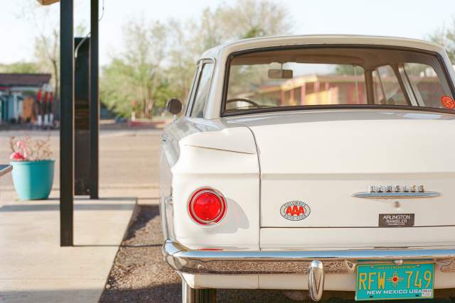 Rear view of a classic white AMC Rambler parked under a shelter with New Mexico license plate and AAA sticker, in a sunny outdoor setting with trees and plants around.