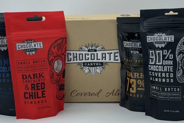 Five packages of Chocolate Cartel products displayed in a row, including varieties such as Dark & Red Chile Almonds, 90% Dark Covered Almonds, and Dark Chocolate Covered Almonds, with a central box featuring the Chocolate Cartel logo.