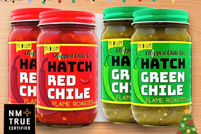 Two jars of Hatch Red Chile and two jars of Hatch Green Chile, labeled "Medium Chop Flame Roasted," displayed with festive lights and a small Christmas tree decoration in the background.