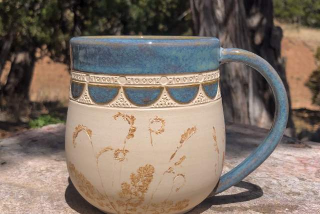 A ceramic mug with a blue rim and handle, featuring a stamped pattern and botanical designs, sits on a stone surface outdoors.