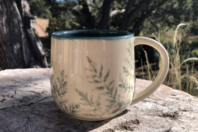 A ceramic mug with a leaf design, sitting on a wooden surface outdoors, with trees in the background.