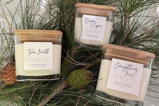 Four scented candles labeled "Tree Wilt," "Sugar & Spice," and "Everything Nice" displayed among pine branches and pine cones on a reflective surface.
