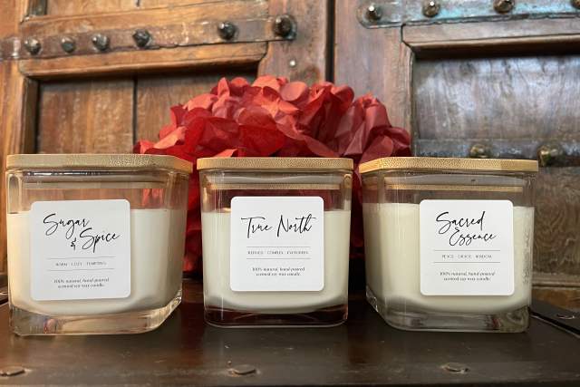 Three scented candles are arranged in a row on a wooden surface against a rustic metal backdrop, accompanied by a bright red floral arrangement in the background.