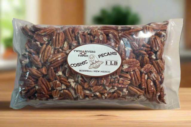 A sealed clear plastic bag filled with pecans, labeled "Twin Rivers Farm Cosmic Pecans 1lb, Roswell, New Mexico," is placed on a wooden surface. A blurred background with green colors suggests a kitchen setting.