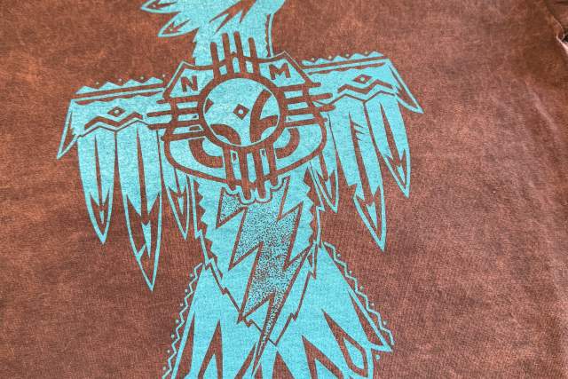 A shirt features a turquoise tribal-style bird design, with stylized wings and tail feathers, along with a Zia symbol imposed over a Route 66 sign.