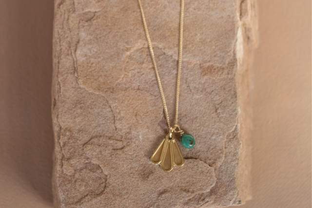 Gold necklace with a fan-shaped pendant and a green bead, hanging on a textured stone surface.