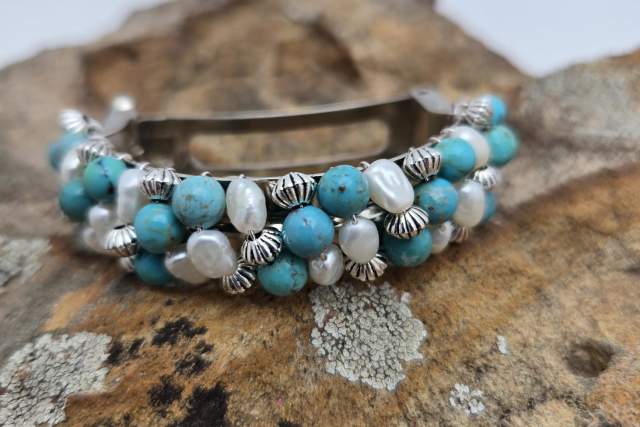 A bracelet with turquoise and pearl beads, interspersed with metal accents, is placed on a textured stone surface.