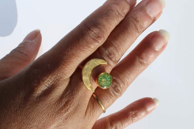 A hand with neatly manicured nails wearing a gold ring featuring a crescent shape and a round, green opal. The ring is positioned on the middle finger against a plain background.