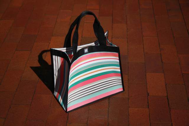 A colorful striped tote bag with black handles is placed on a red brick pavement, casting a shadow.