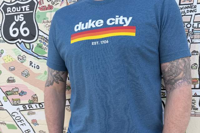 A person wearing a colorful "duke city" t-shirt stands in front of a colorful mural featuring Route 66 and various map elements.