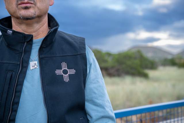 Close-up of a person wearing a vest with the Zia sun symbol, standing outdoors with a blurred background of desert landscape.