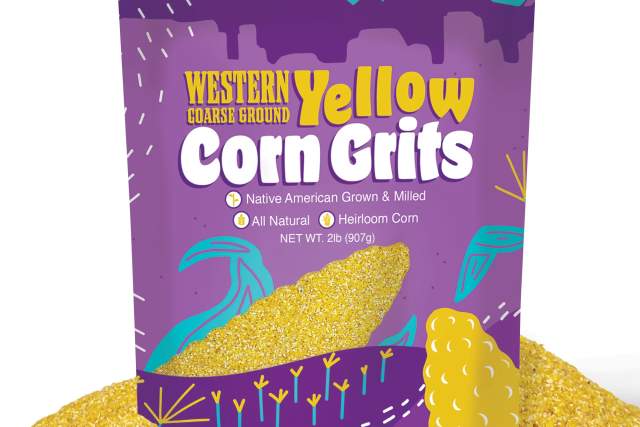 The image shows a colorful package of "Western Coarse Ground Yellow Corn Grits." The packaging features a vibrant design with mountains, corn kernels, and text highlighting it as "Native American Grown & Milled," "All Natural," and "Heirloom Corn." A pile of yellow corn grits is seen spilling out in front of the package.