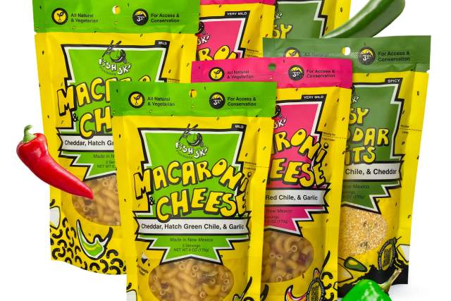 Variety of FishSki Provisions Macaroni and Cheese packages, including flavors like Cheddar, Hatch Green Chile, Red Chile & Garlic, displayed with fresh peppers.