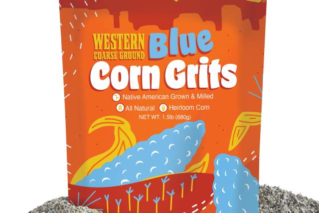 A package of Western Blue Corn Grits, featuring bright, colorful graphics with depictions of corn and landscapes. The packaging highlights that the product is Native American grown and milled, all-natural, and made from heirloom corn. The bag sits on a small pile of gray coarse ground material.