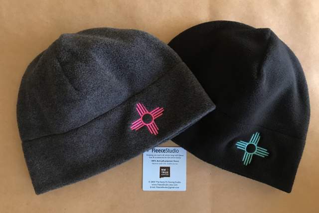 Two fleece beanies displayed side-by-side; one in gray and the other in black, both featuring a Zia sun symbol, positioned above a label from FleeceStudio.