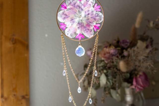 Decorative hanging ornament featuring a circular resin piece with encapsulated pink flowers, adorned with multiple dangling chains and teardrop crystals.
