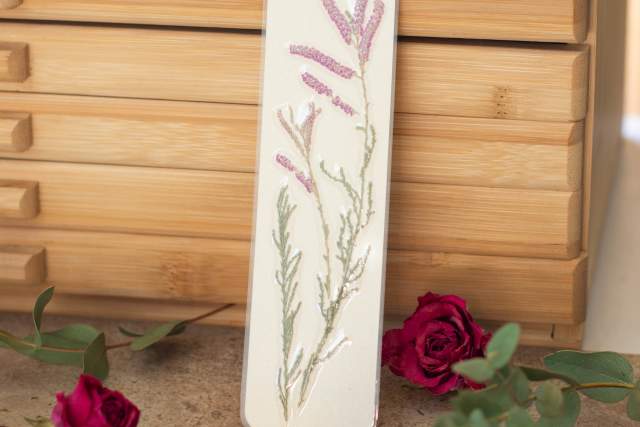 A floral bookmark with a design of purple lavender and green stems rests against a light wooden box, surrounded by deep red roses and green leaves.
