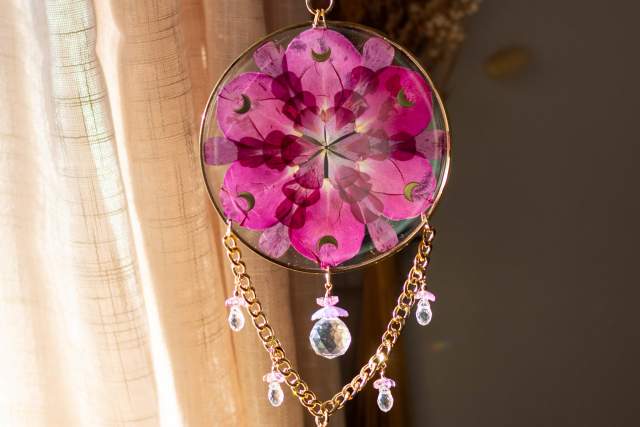 A decorative hanging featuring a round frame with pressed flowers and a kaleidoscope-like design. It is adorned with dangling crystal-like beads and chains, capturing sunlight and casting colorful reflections.