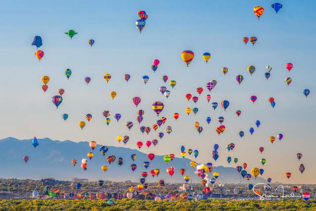 Colorful hot air balloons fill the sky at the Albuquerque International Balloon Fiesta, with distant mountains under a clear blue sky.