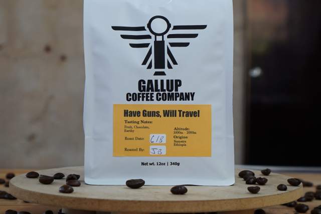 A paper package of coffee labeled "Gallup Coffee Company" with the text "Have Guns, Will Travel" sits on a round wooden platform. Coffee beans are scattered around the platform.