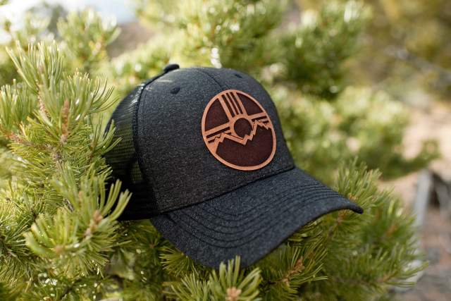 Black baseball cap with a leather patch featuring a Zia symbol behind a mountain, nestled in a pine tree branch.