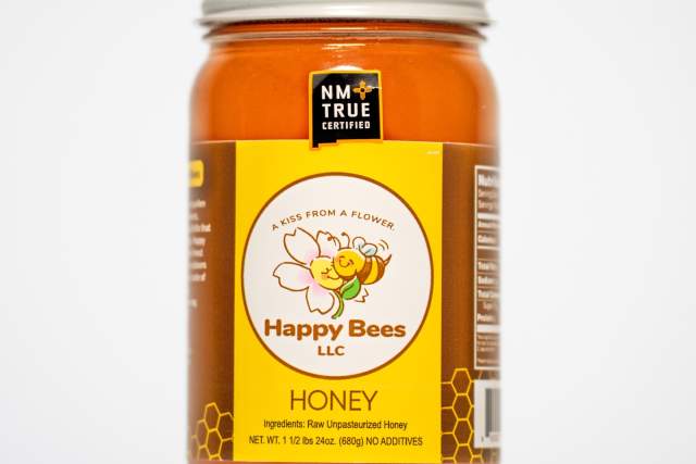 Jar of "Happy Bees LLC" raw, unpasteurized honey with label featuring a cartoon bee, certified by "NM True".