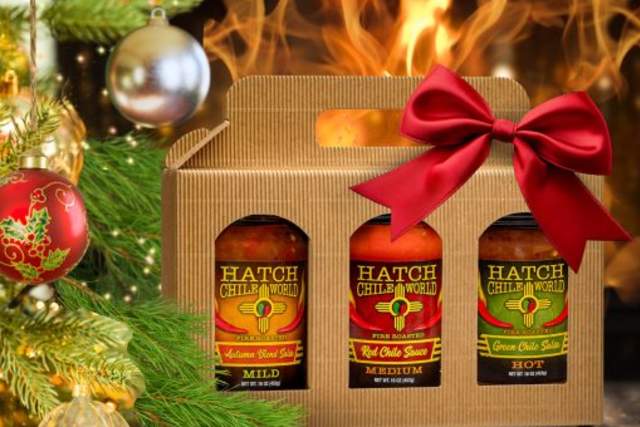 Gift box containing three bottles of Hatch Chile World sauces with mild, medium, and hot varieties, decorated with a red bow, placed beside a Christmas tree ornament and in front of a cozy fireplace.
