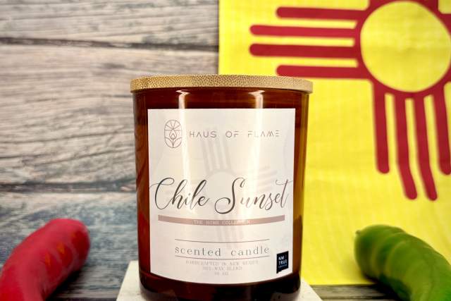 A scented candle labeled "Chile Sunset" by Haus of Flame, placed on a small stand, with a decorative yellow background featuring a red Zia symbol. Two chili peppers are adjacent to the candle.