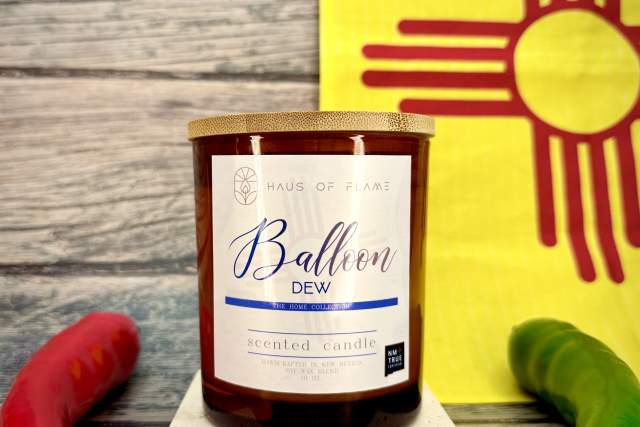 A scented candle branded "Haus of Flame" with the scent name "Balloon Dew" on a wooden surface, flanked by a red and a green chili pepper, with a yellow sign featuring the Zia sun symbol in the background.