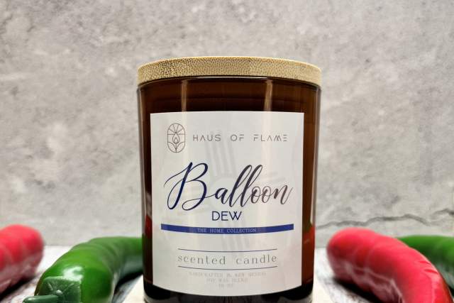A scented candle labeled "Balloon Dew" from Haus of Flame, surrounded by red and green chili peppers on a textured surface.